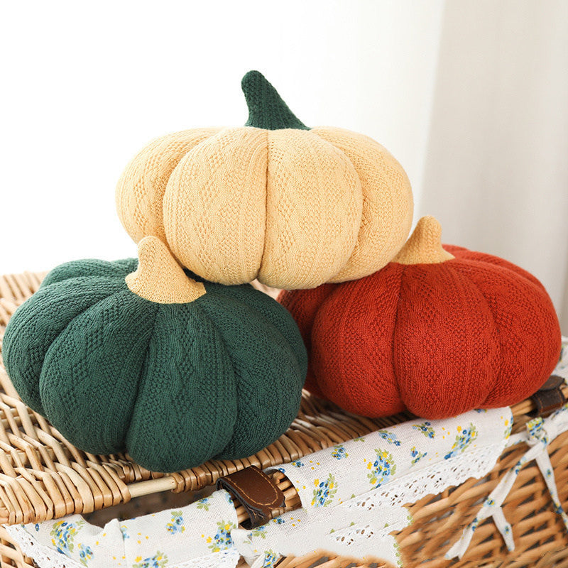 Cable-knit plush pumpkin pillows in butter, forest green and rust stacked on a wicker chest